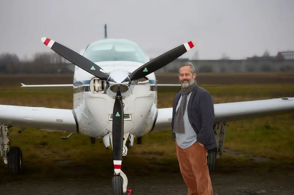 Ein Mann mit grauem Haar und Bart steht lächelnd vor einem kleinen weißen Propellerflugzeug an einem bewölkten Tag, mit Gras und Bäumen im Hintergrund.