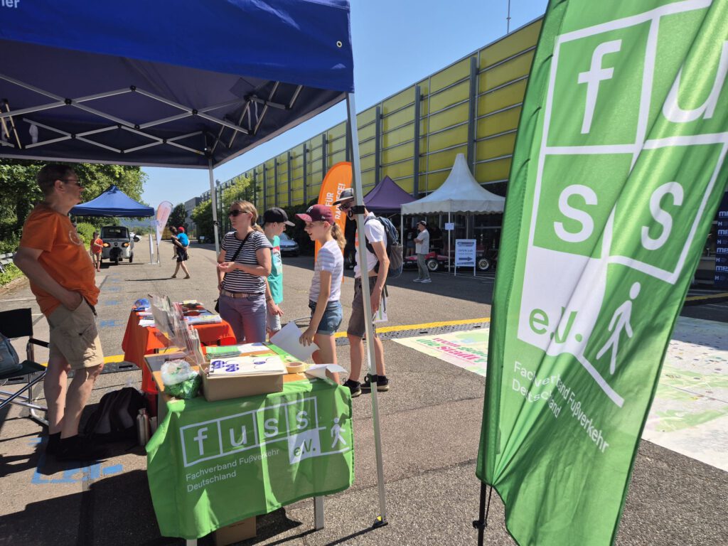 Unter einem blauen Vordach stehen Menschen an einem Stand im Freien, auf dem Informationsmaterial ausliegt. Im Vordergrund ist eine große grüne Fahne mit der Aufschrift „fuss e.V.“ und einem Fußgängersymbol zu sehen. Im Hintergrund sind weitere Stände und Banner zu erkennen.