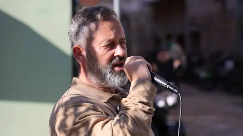 Man with gray beard singing into a handheld microphone outdoors, wearing a beige shirt.