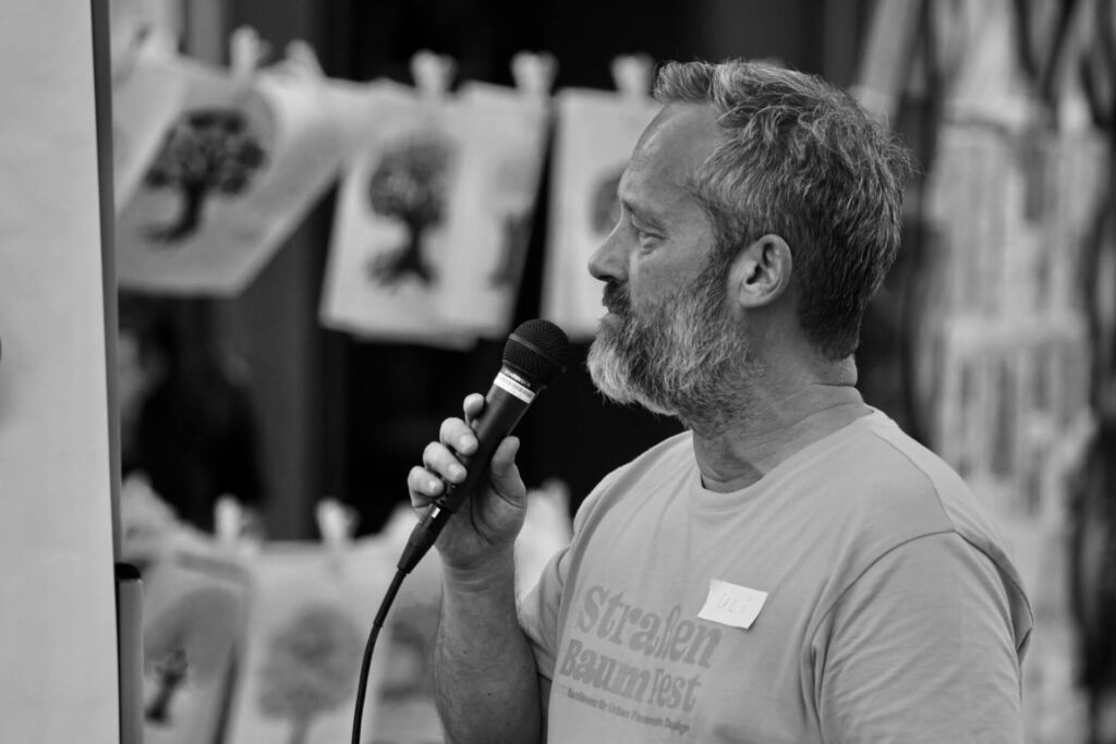 Ein bärtiger Mann spricht bei einer Veranstaltung im Freien in ein Mikrofon, hinter ihm hängen Zeichnungen von Bäumen. Er trägt ein T-Shirt und ein Namensschild und blickt auf dem Foto nach links. Das Bild ist in Schwarz-Weiß.