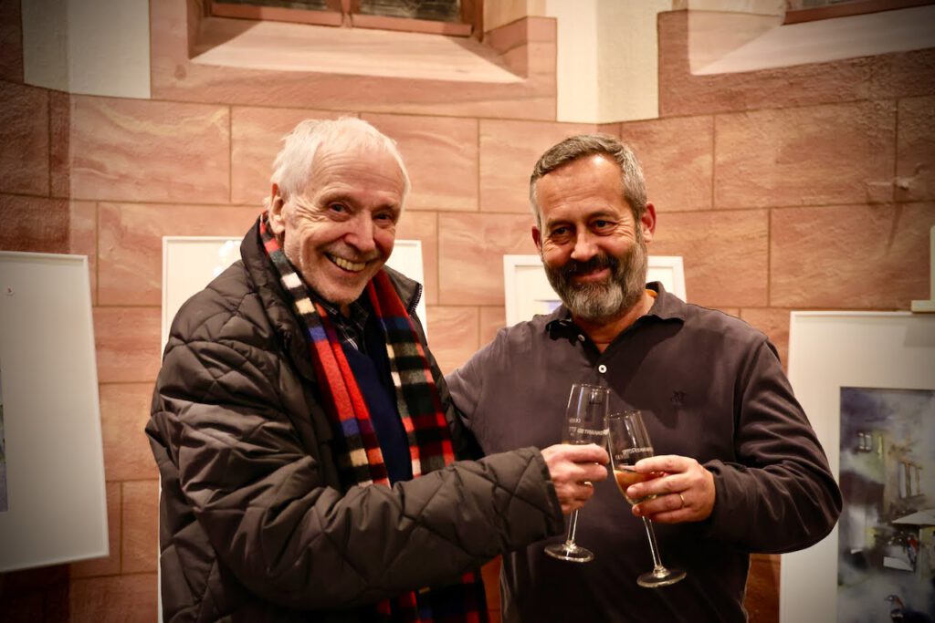 Zwei lächelnde Männer stoßen in einem warm beleuchteten Raum, an dessen Wänden gerahmte Kunstwerke hängen, mit ihren Gläsern Weißwein an; der eine trägt einen bunten Schal und eine Jacke, der andere ein dunkles Hemd.