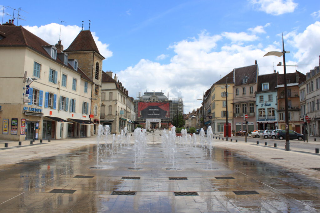 Ein Stadtplatz mit Springbrunnen im Vordergrund, umgeben von historischen europäischen Gebäuden, Geschäften und geparkten Autos unter einem teilweise bewölkten Himmel.