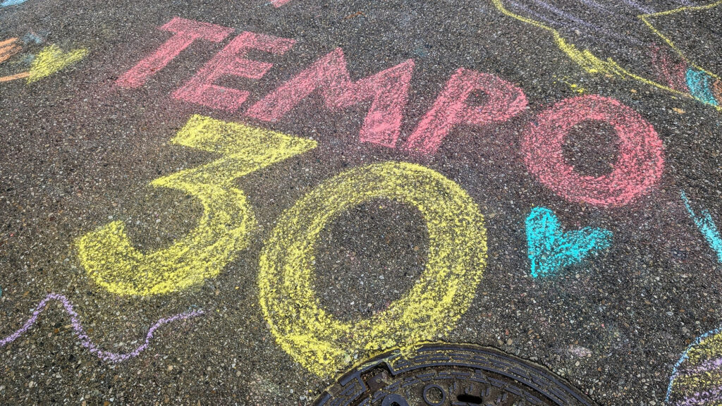 Eine farbenfrohe Kreidezeichnung auf dem Bürgersteig, auf der in großen Buchstaben und Zahlen „TEMPO 30“ zu lesen ist, daneben sind Herzen und Schnörkel zu sehen. In der rechten unteren Ecke ist ein Kanaldeckel zu erkennen.