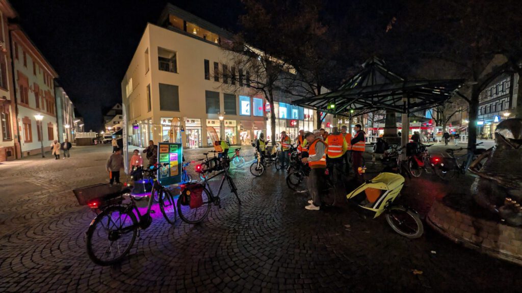 Group of people in high-visibility vests with bicycles under a glass canopy in a cobblestone town square at night.