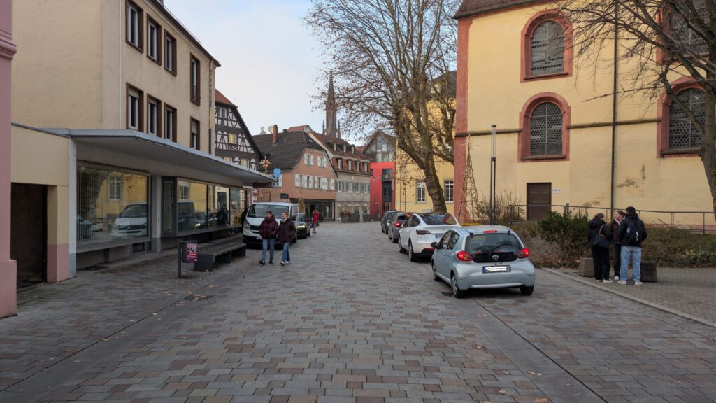 Eine Kopfsteinpflasterstraße mit geparkten Autos, Menschen, die spazieren gehen und sich unterhalten, sowie historischen europäischen Gebäuden, vor denen an einem bewölkten Tag Bäume den Bürgersteig säumen.