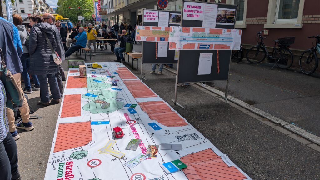 Menschen versammeln sich um eine große Karte, die auf der Straße ausgebreitet ist; daneben stehen Informationstafeln und Plakate. Tische, Fahrräder und Gebäude säumen das Stadtbild und lassen auf eine Gemeinschaftsveranstaltung oder eine Diskussion über Stadtplanung schließen.