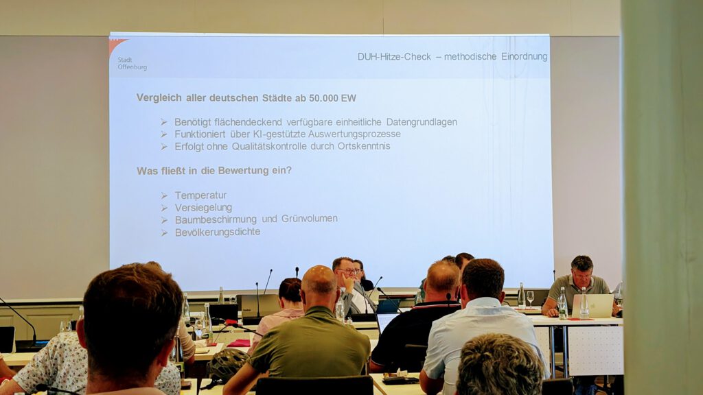 Eine Gruppe von Menschen sitzt in einem Konferenzraum und verfolgt eine projizierte Präsentation. Die Folie befasst sich mit einer Methode zur Hitzebelastungsanalyse für deutsche Städte und listet Kriterien und Bewertungsfaktoren wie Temperatur und Bevölkerungsdichte auf.