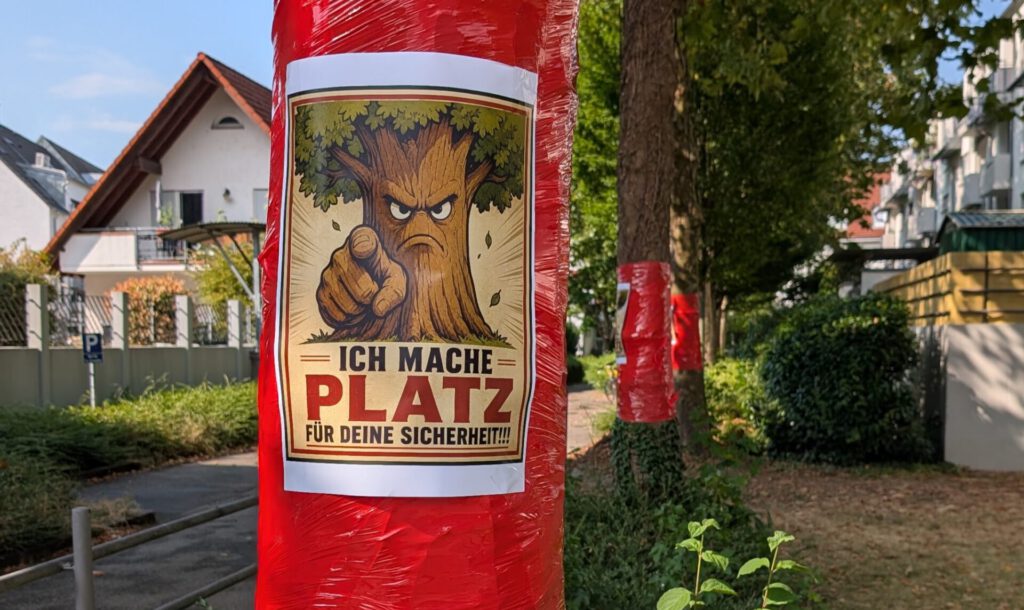 An einem mit roter Plastikfolie umwickelten Baum entlang einer Wohnstraße ist ein Plakat mit der Abbildung eines wütenden Comic-Baums angebracht, der mit dem Finger zeigt, sowie dem deutschen Text „Ich mache Platz für deine Sicherheit!!!“.