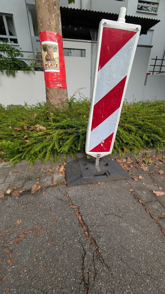 Ein rot-weißes Baustellenschild steht auf einem rissigen Bürgersteig neben grünen Büschen und einem Baum. Um den Baumstamm ist ein Plakat mit einem Hund und einem deutschen Text gewickelt.