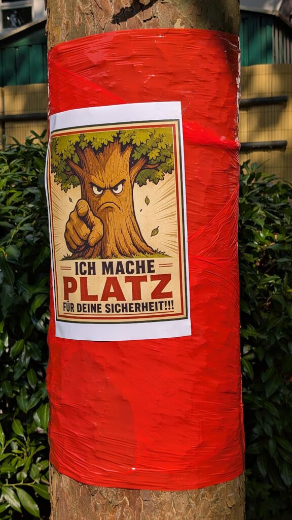 An einem mit rotem Klebeband umwickelten Baumstamm ist ein Plakat mit der Abbildung eines wütenden Comic-Baums zu sehen, der mit dem Finger zeigt, sowie der Text auf Deutsch: „Ich mache Platz für deine Sicherheit!!!“ Im Hintergrund sind Büsche und ein Zaun zu erkennen.