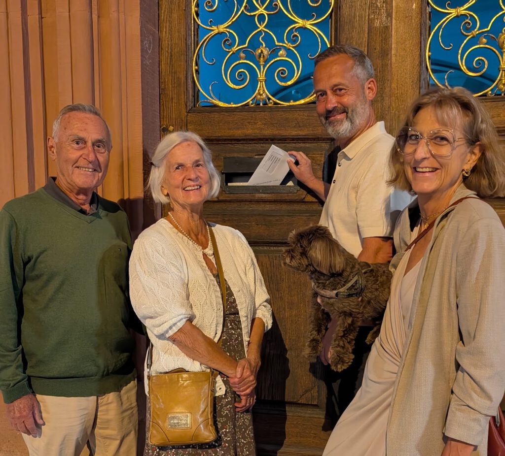 Group of four seniors posing outside a wooden door with gold decorative ironwork; a small brown dog is held by one person.