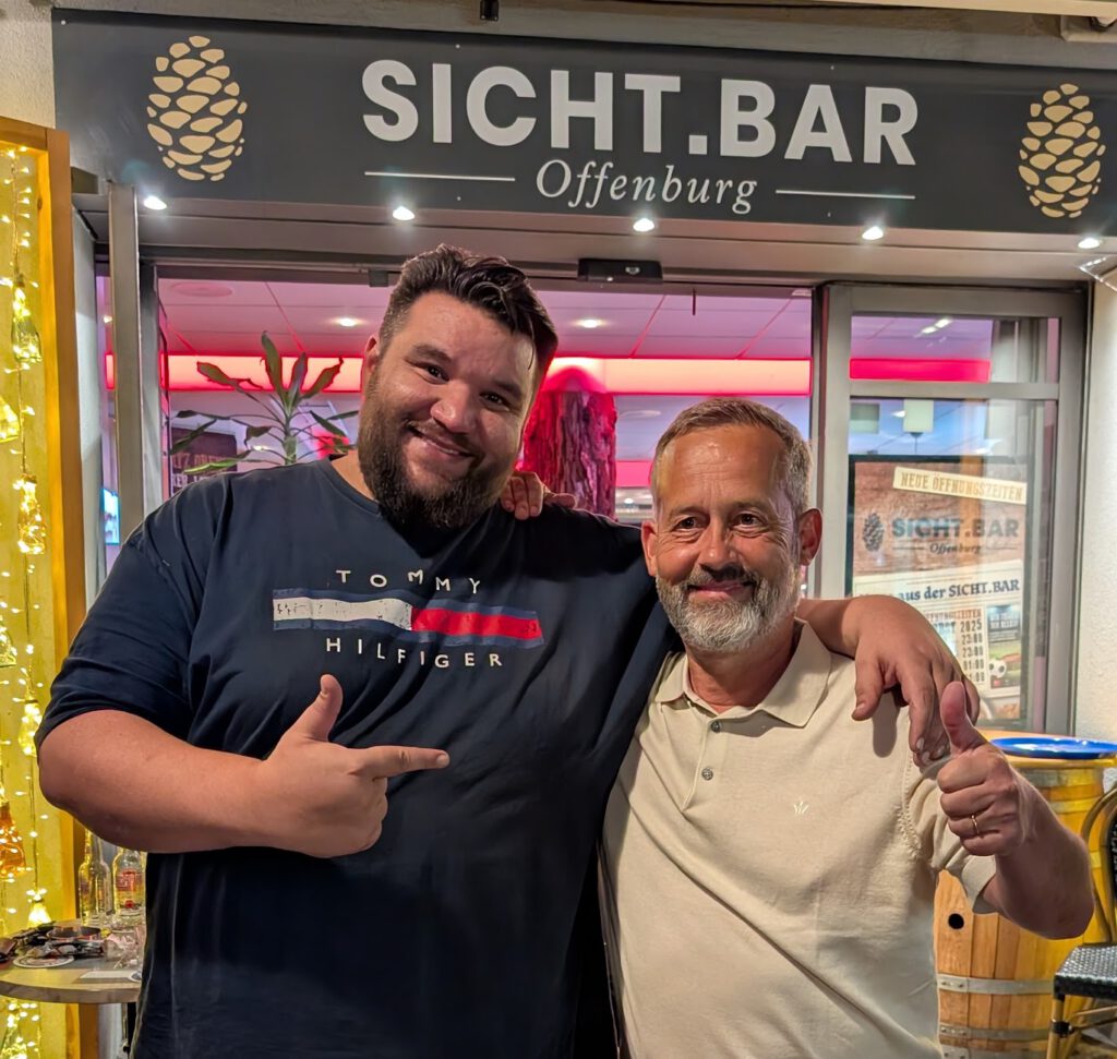 Two men smile with arms around each other outside Sicht.Bar Offenburg, posing for the photo.