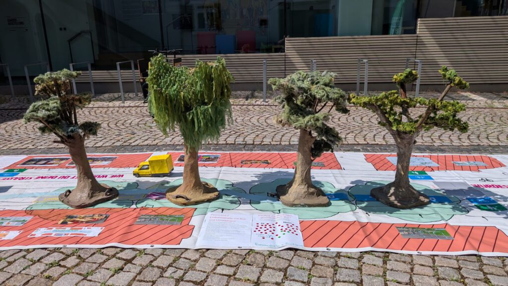Vier Modellbäume stehen auf einer bunten Lernmatte mit Abbildungen und Text, die im Freien auf Kopfsteinpflaster liegt; neben dem Baum ganz links steht ein kleiner gelber Lastwagen.