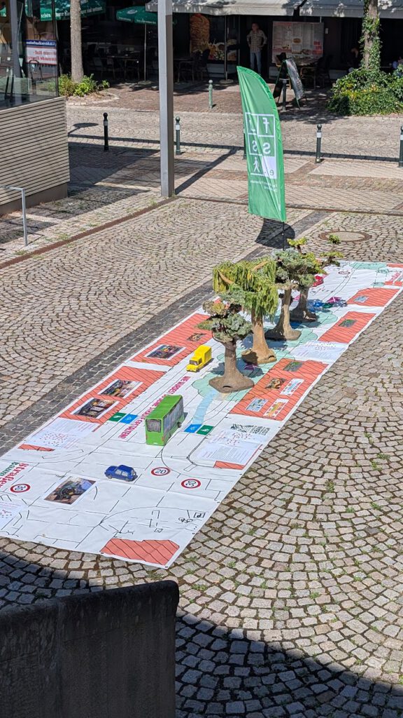 Auf einem mit Kopfsteinpflaster gepflasterten Platz ist ein großes Spielbrett mit Spielzeugautos und Modellbäumen aufgebaut. Daneben weht eine grüne Flagge, und im Hintergrund stehen Tische, an denen Menschen sitzen.