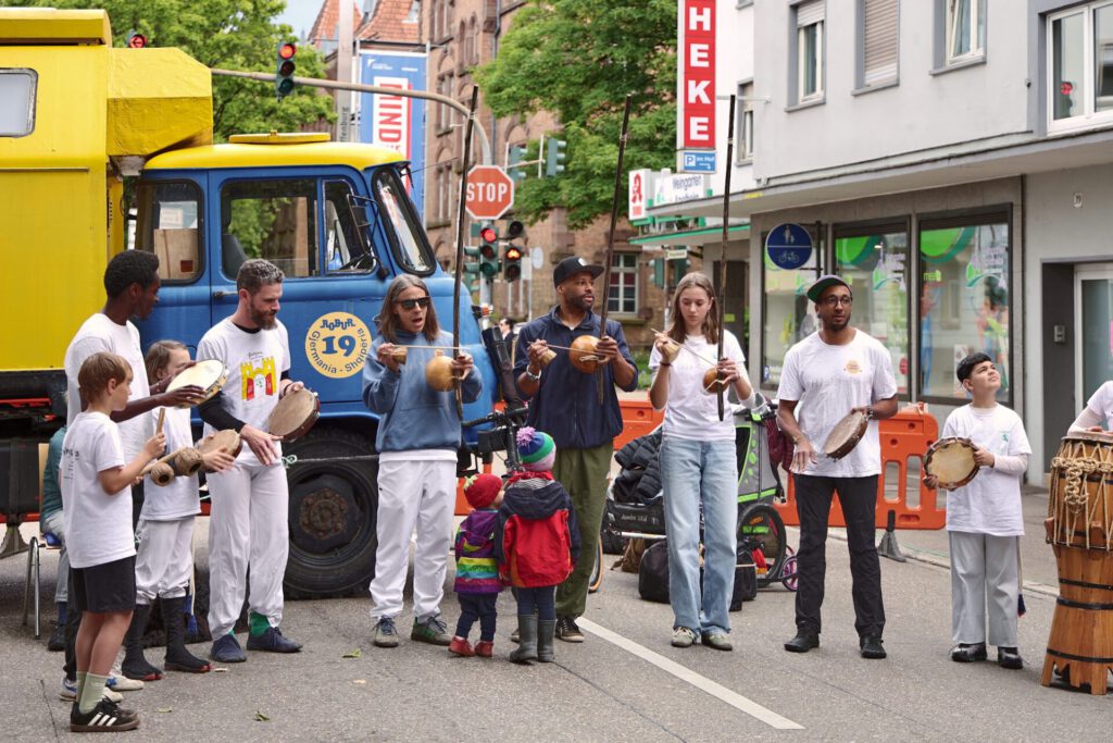 Eine bunte Gruppe von Menschen, darunter Kinder und Erwachsene, spielt auf einer Straße Percussion-Instrumente, im Hintergrund sind ein gelb-blauer Lkw und orangefarbene Absperrungen zu sehen. Einige Kinder schauen zu, während andere mitspielen.