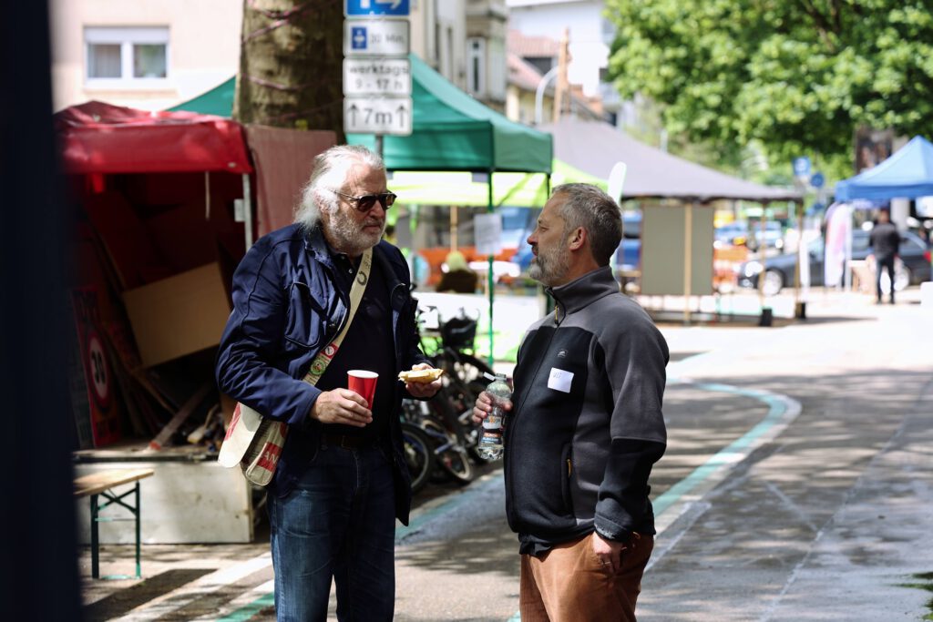 Zwei Männer stehen auf einer Straße, die von Zelten und Marktständen gesäumt ist, und unterhalten sich. Der eine hält einen roten Becher und etwas zu essen in der Hand, der andere eine Wasserflasche. Es scheint sich um eine Veranstaltung im Freien oder einen Markt an einem sonnigen Tag zu handeln.