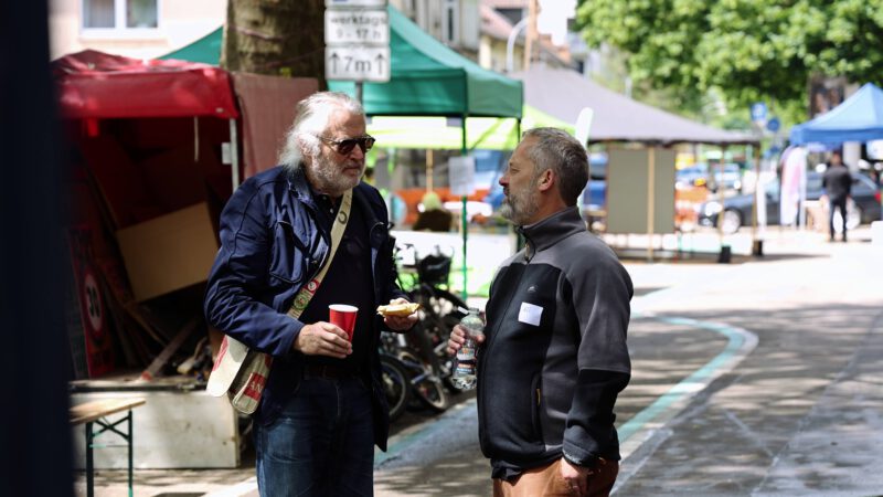 Zwei Männer stehen auf einer Straße, die von Zelten und Marktständen gesäumt ist, und unterhalten sich. Der eine hält einen roten Becher und etwas zu essen in der Hand, der andere eine Wasserflasche. Es scheint sich um eine Veranstaltung im Freien oder einen Markt an einem sonnigen Tag zu handeln.