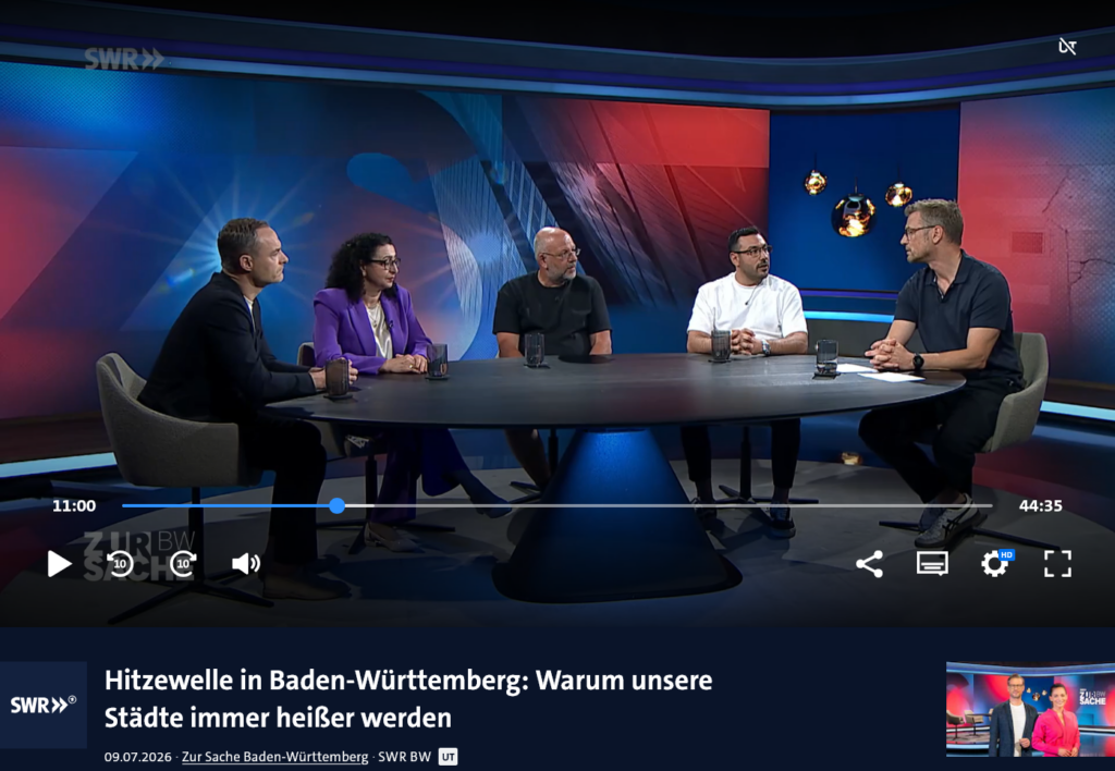 Fünf Personen sitzen in einem Fernsehstudio um einen runden Tisch herum und diskutieren. Auf dem Bildschirm ist ein deutscher Text über Hitzewellen in Baden-Württemberg zu sehen und darüber, warum es in den Städten immer heißer wird.