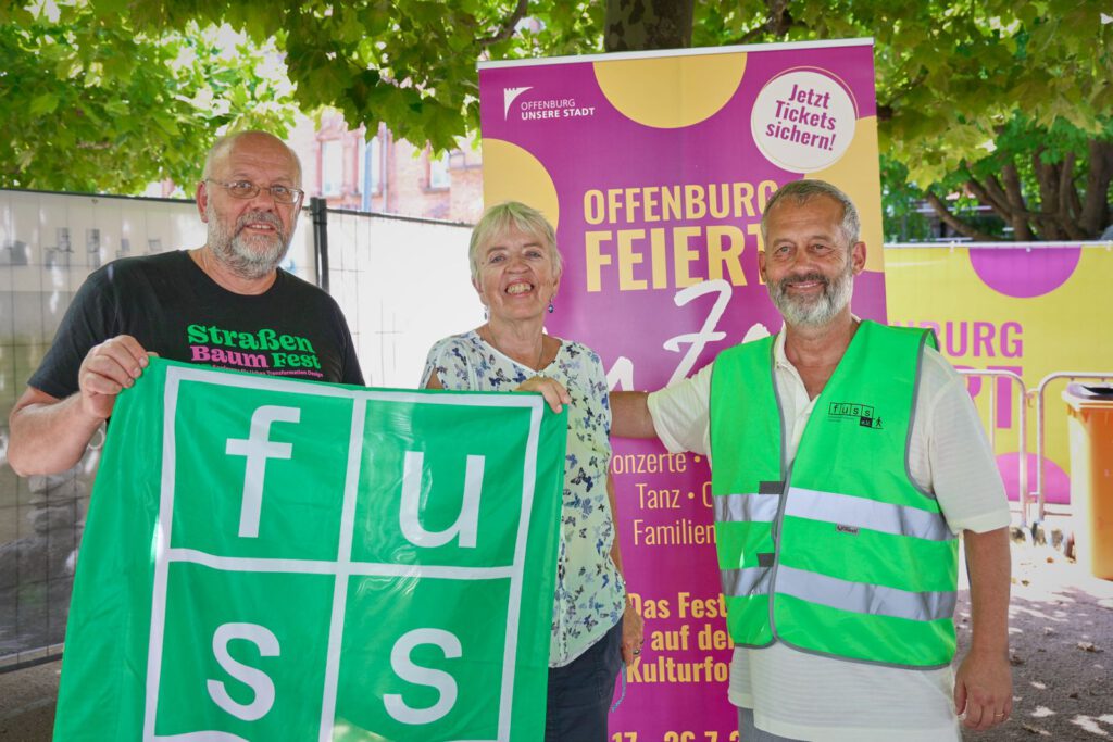 Drei lächelnde Erwachsene stehen im Freien vor einem Festivalbanner. Zwei Männer halten eine grüne „Fuss“-Fahne in den Händen, einer trägt eine grüne Weste. Eine Frau in der Mitte trägt ein geblümtes Hemd. Im Hintergrund sind Bäume und bunte Schilder zu sehen.