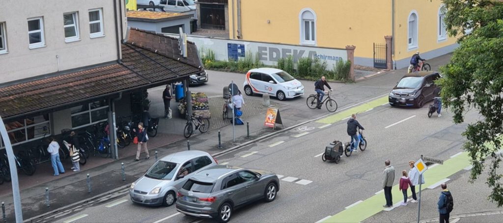 Eine belebte Straßenszene mit Autos, Radfahrern, Fußgängern und Menschen, die vor einem Fahrradgeschäft stehen. Einige überqueren die Straße, andere schieben Einkaufswagen oder gehen mit Hunden spazieren. Im Hintergrund sind ein gelbes Gebäude und ein Lebensmittelgeschäft zu sehen.