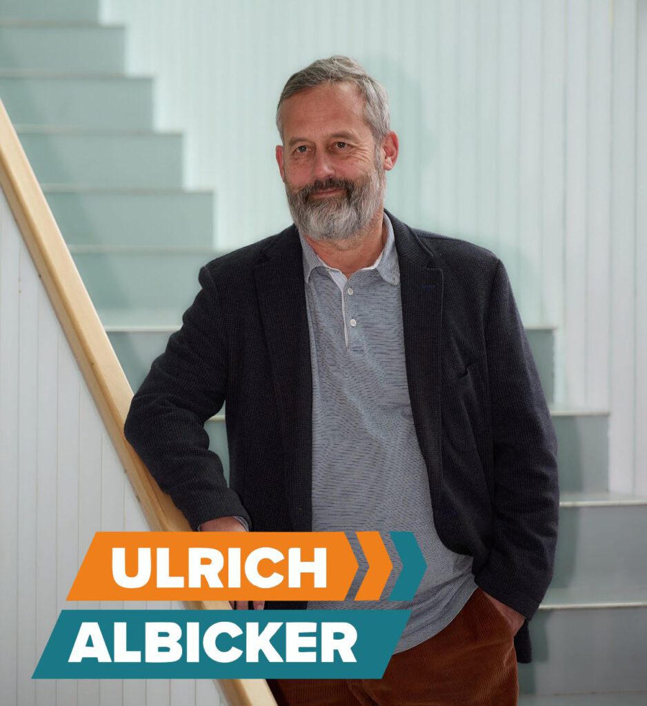 Ein Mann mit grauen Haaren und Bart steht auf einer Treppe und trägt ein graues Hemd und einen dunklen Blazer. Vor ihm ist in fetten orangefarbenen, weißen und türkisfarbenen Buchstaben der Schriftzug „ULRICH ALBICKER“ zu lesen.