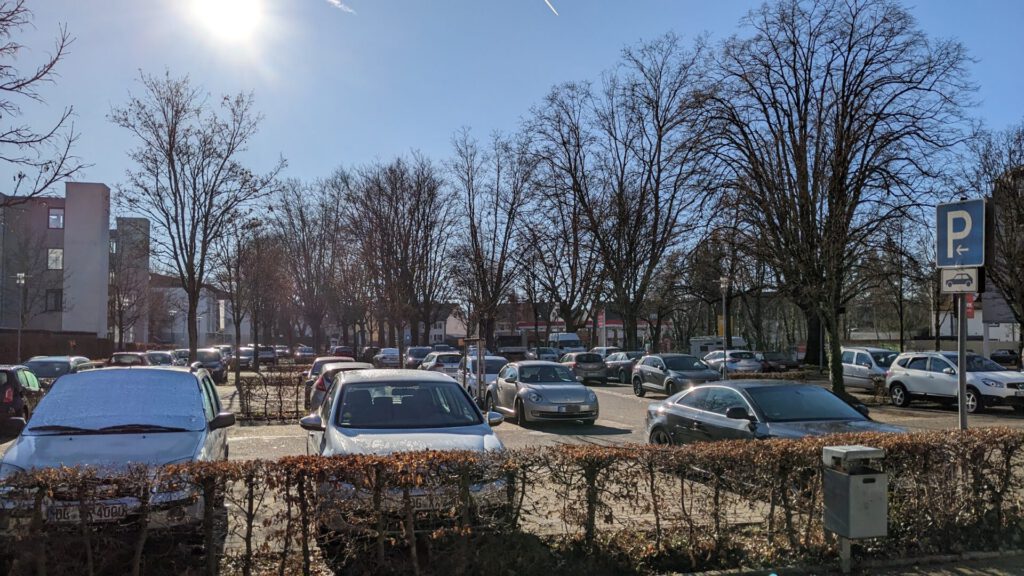 Ein sonniger Tag auf einem Parkplatz mit vielen geparkten Autos, kahlen Bäumen, einer Hecke im Vordergrund und einem blauen Parkschild auf der rechten Seite. Das Sonnenlicht wirft Schatten über die Szene.