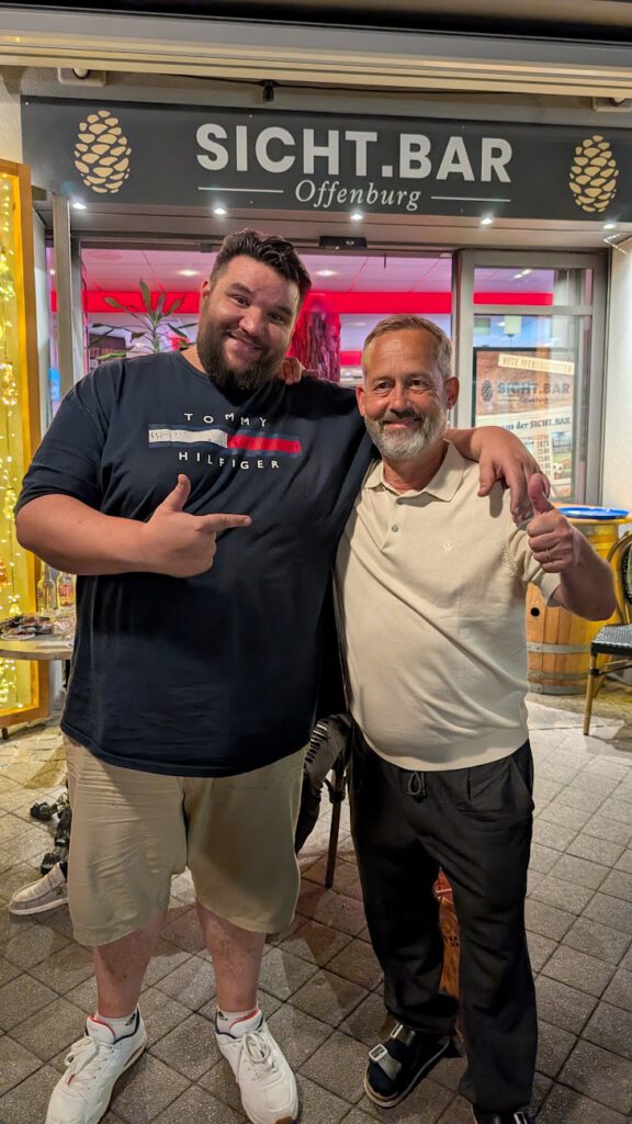 Rezan und Uli. Zwei lächelnde Männer posieren nachts gemeinsam vor der Sicht-Bar in Offenburg. Ein Mann zeigt auf den anderen. Hinter ihnen sind große Fenster und das beleuchtete Schild der Bar zu sehen.