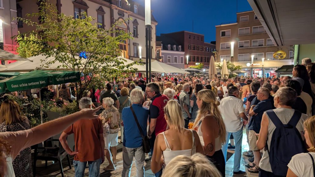 Nachts versammelt sich eine große Menschenmenge im Freien auf einem belebten Stadtplatz, umgeben von Gebäuden und Sonnenschirmen. Die Menschen stehen herum, unterhalten sich und genießen die festliche Atmosphäre im Schein der Straßenlaternen.
