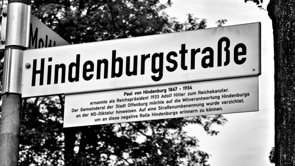 Ein Straßenschild mit der Aufschrift „Hindenburgstraße“ und einer Gedenktafel, die den historischen Zusammenhang zu Paul von Hindenburg und Adolf Hitler erläutert, fotografiert in Schwarz-Weiß vor dem Hintergrund von Bäumen.
