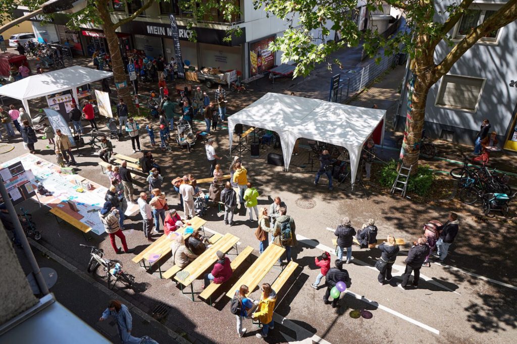 Ein Straßenfest im Freien, bei dem sich die Menschen um Tische und Zelte versammelt haben – manche sitzen, manche stehen. Bäume spenden teilweise Schatten. Stände und Auslagen säumen die Straße. Die Atmosphäre wirkt lebhaft und gesellig.