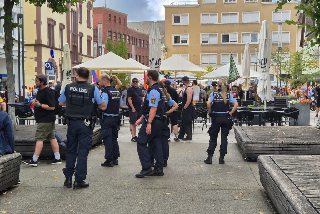 Mehrere Polizeibeamte stehen auf einem öffentlichen Platz in der Nähe von Sitzgelegenheiten im Freien, während sich Menschen um sie herum versammeln. Einige Personen unterhalten sich mit den Beamten. Im Hintergrund sind umliegende Gebäude und Sonnenschirme zu sehen.