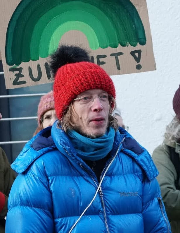 Fritz Düker mit einer roten Strickmütze und einer blauen Jacke steht im Freien bei einer Demonstration, im Hintergrund ist ein Schild mit einem grünen Regenbogen und teilweise lesbarem deutschen Text zu sehen.