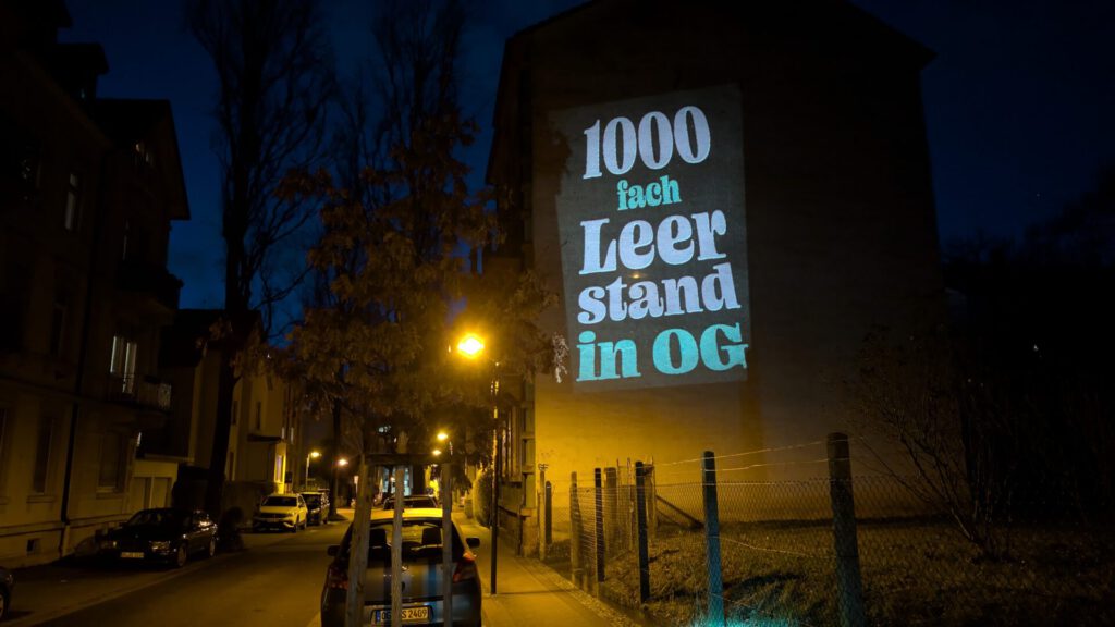 Eine nächtliche Straßenszene mit einer Textprojektion an einem Gebäude, auf der „1000 fach Leer stand in OG“ zu lesen ist. Geparkte Autos und Straßenlaternen säumen die Straße; kahle Bäume stehen am Straßenrand.