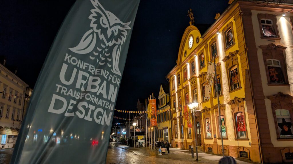Eine nächtliche Straßenszene zeigt ein Banner mit einem Eulenlogo und der Aufschrift „Konferenz für Urban Transformation Design“ neben einem beleuchteten historischen Gebäude, an dessen Fassade Kunstwerke ausgestellt sind.