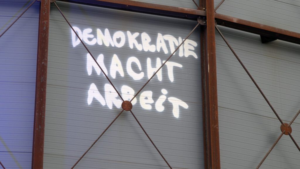 Auf einer Wellblechfläche ist in weißer Graffiti-Schrift „DEMOKRATIE MACHT ARBEIT“ zu lesen. Die Wand ist von rostigen Metallträgern und Querstreben eingerahmt.