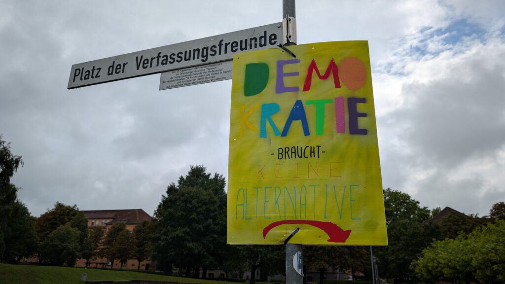 Ein buntes Schild mit der Aufschrift „Demokratie braucht keine Alternative“ hängt unter einem Straßenschild mit der Bezeichnung „Platz der Verfassungsfreunde“ in einem Park, im Hintergrund sind Bäume und Gebäude zu sehen.