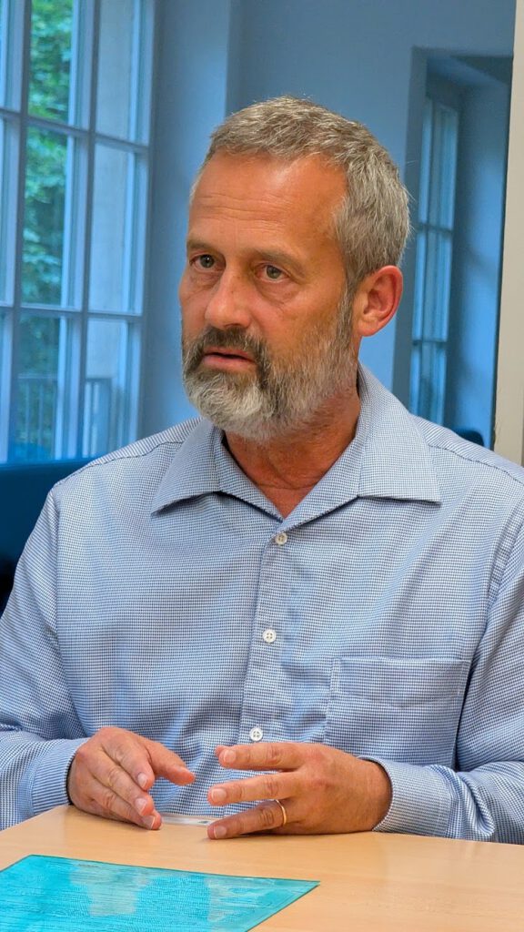 Uli Albicker mit grauen Haaren und Bart, der ein hellblaues kariertes Hemd trägt, sitzt an einem Schreibtisch, vor sich ein türkisfarbenes Blatt Papier, und gestikuliert mit den Händen in einem hell beleuchteten Raum mit großen Fenstern.
