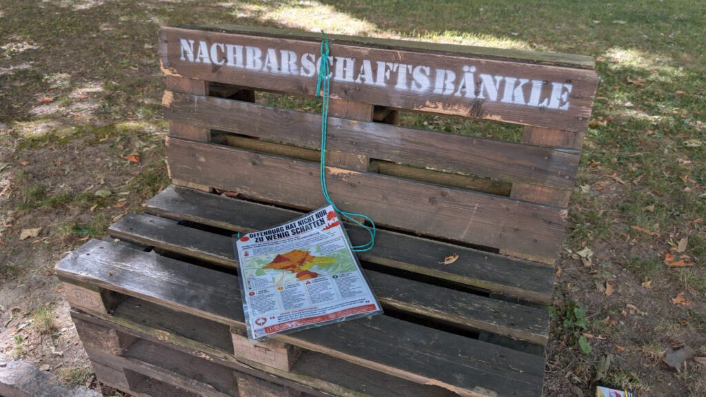 Auf einer Holzbank mit der Aufschrift „NACHBARSCHAFTSBÄNKLE“ liegt ein grünes Seil drüber, und auf der Sitzfläche liegt eine Zeitung mit einer Karte. Die Bank steht im Freien auf Gras und trockenen Blättern.