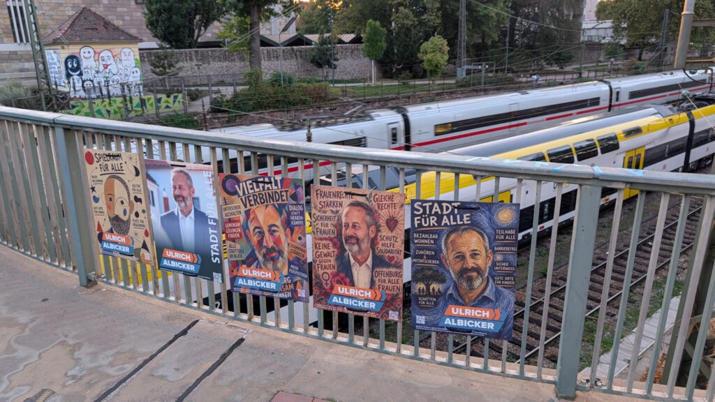 Auf einer Brückenbrüstung mit Blick auf eine Bahnstrecke, auf der gerade ein Zug vorbeifährt, sind Wahlplakate zu sehen, auf denen ein Kandidat mit Bart abgebildet ist; im Hintergrund sind Graffiti und Bäume zu erkennen.