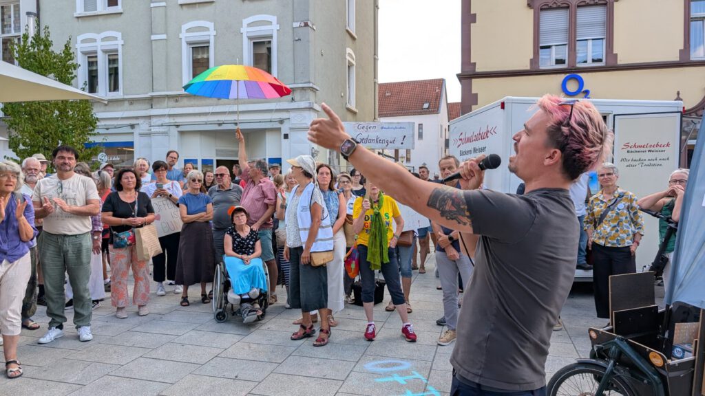 Eine Person mit einem Mikrofon spricht auf einem Platz im Freien zu einer bunt gemischten Menschenmenge; ein Teilnehmer hält einen Regenbogenschirm in der Hand, und viele Menschen hören aufmerksam zu.