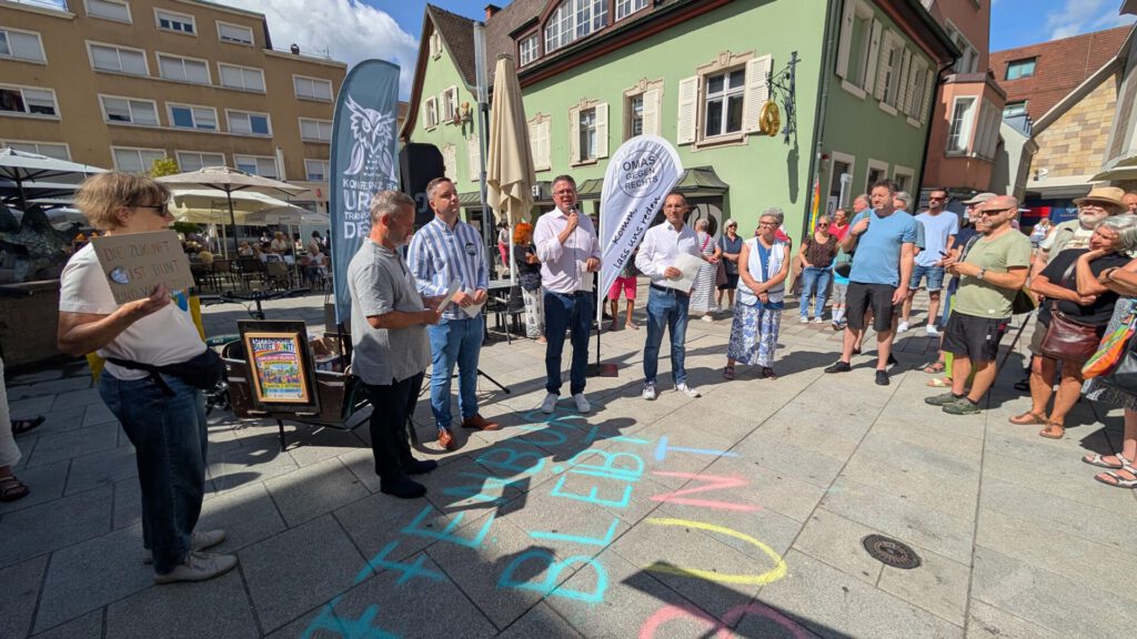 Eine Gruppe von Menschen steht draußen auf einem Platz, einige halten Schilder und Transparente in den Händen. Auf dem Boden sind bunte Kreideinschriften zu sehen, und mehrere Schaulustige beobachten die Szene an einem sonnigen Tag.