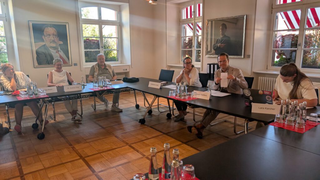Sechs Personen sitzen an Tischen in einem Besprechungsraum mit großen Fenstern und Porträts an den Wänden. Einige unterhalten sich, andere hören zu oder schreiben. Auf den Tischen stehen Wasserflaschen und liegen Papiere.