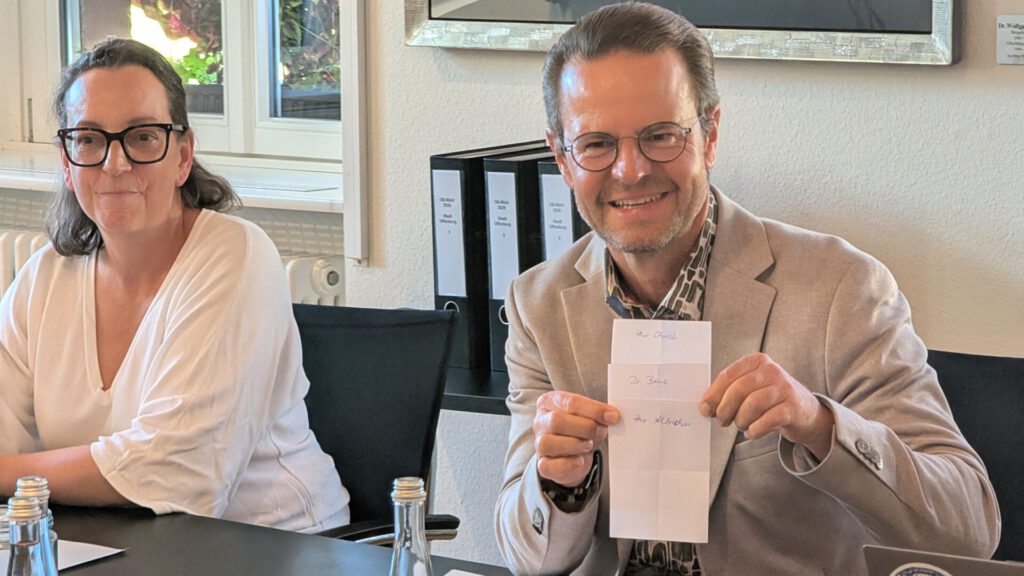 Ein Mann mit Brille und einem beigen Blazer lächelt, während er ein gefaltetes Blatt Papier in der Hand hält. Eine Frau mit Brille und einem weißen Oberteil sitzt neben ihm an einem Tisch, auf dem Wasserflaschen stehen. Im Hintergrund sind Büroordner und Kunstwerke zu sehen.