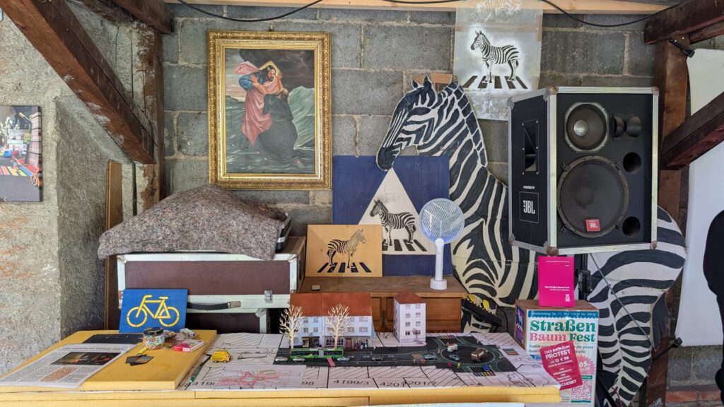 Ein überfüllter Raum mit Zebra-Dekorationen, einem Gemälde, einem Lautsprecher, einer Fahrrad-Schablone, Landkarten und verschiedenen Papiergegenständen, die auf einem Tisch vor einer Backsteinwand angeordnet sind.