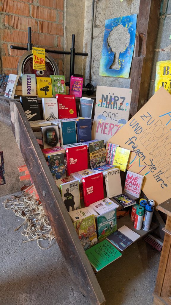 In einem rustikalen Raum mit Ziegel- und Betonwänden sind bunte Bücher und Plakate auf einem Holzständer ausgestellt. Kunstwerke, Sprühdosen und handgeschriebene Schilder tragen zur kreativen, ungezwungenen Atmosphäre bei.