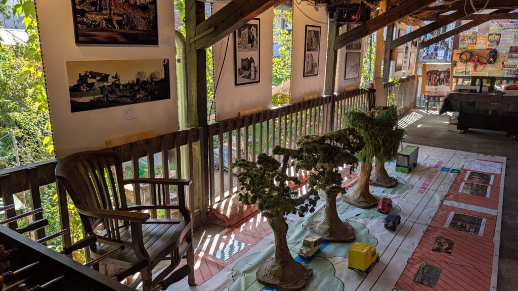 Eine überdachte Veranda ist mit Holzstühlen, gerahmten Fotos an den Wänden und einer Ausstellung auf dem Boden mit Modellbäumen und kleinen Gegenständen ausgestattet, die von natürlichem Sonnenlicht beleuchtet werden.