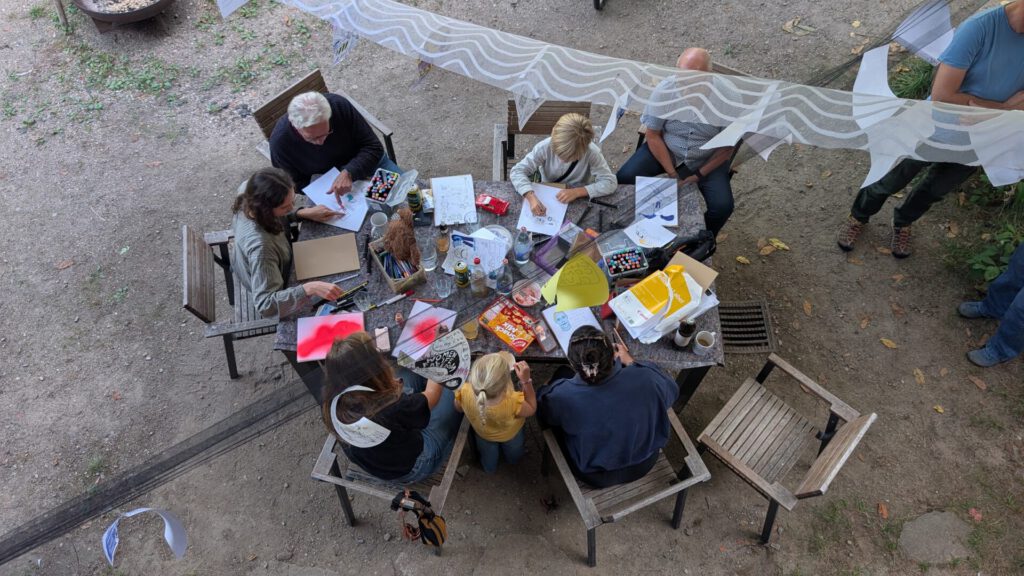 Sieben Personen sitzen im Freien um einen Tisch herum und beschäftigen sich mit Zeichnen oder Basteln, wobei Mal- und Bastelmaterialien auf dem Tisch verstreut liegen. Einige konzentrieren sich auf ihre Arbeit, während andere sich offenbar unterhalten.