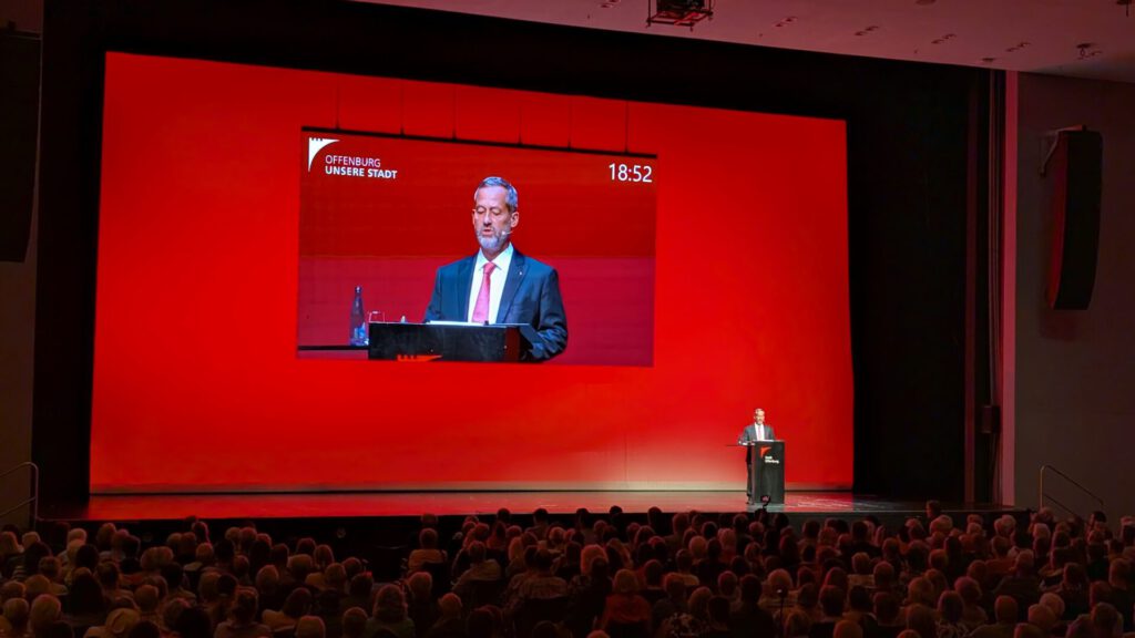Ein Mann im Anzug spricht an einem Rednerpult auf einer Bühne mit rotem Hintergrund, während sein Bild auf eine große Leinwand über ihm projiziert wird. Das Publikum verfolgt die Szene von den Sitzplätzen aus.
