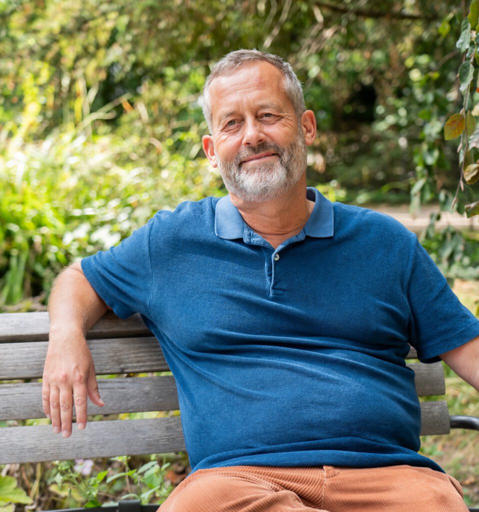 Ein älterer Mann mit grauem Bart und blauem Polohemd sitzt draußen auf einer Holzbank und lächelt leicht. Im Hintergrund sind grüne Bäume und Pflanzen zu sehen.