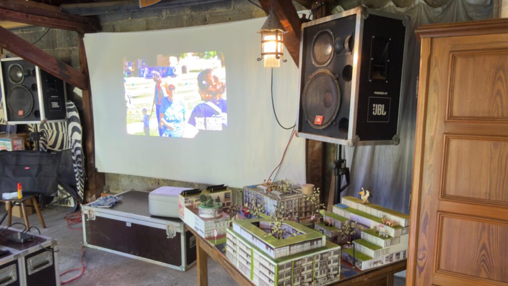 Ein Projektor projiziert eine Außenaufnahme auf eine Leinwand in einem rustikalen Raum, der mit Lautsprechern, Möbeln und einem Tisch voller Miniaturgebäude und Figuren ausgestattet ist, was an eine Spielanordnung für ein Tischspiel erinnert.